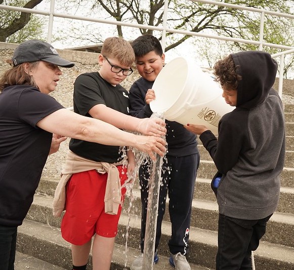 Ginny Carlton assists three students while they pour water into a tall plastic tube