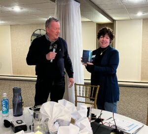 Two people standing next to each other in a room. One person is holding a microphone. The other person is holding an award.