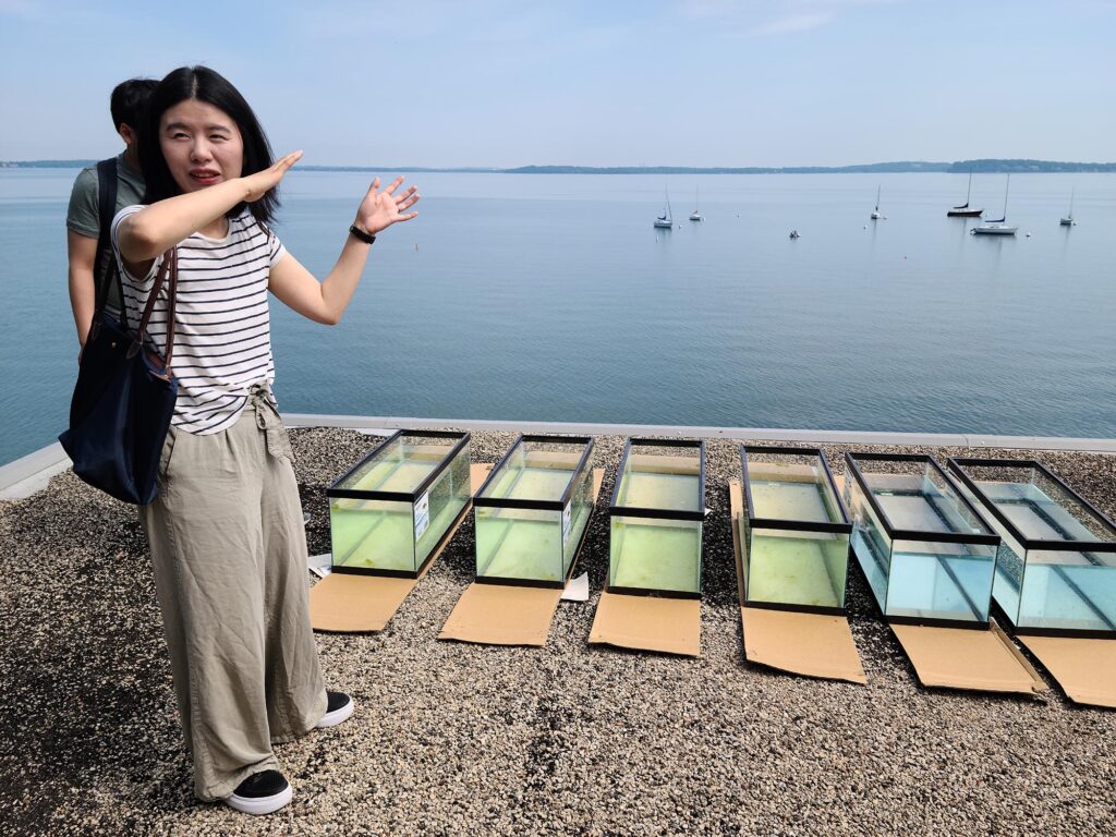 Mohan Qin stands next to six aquariums filled with water with a view a Lake Mendota in the background.