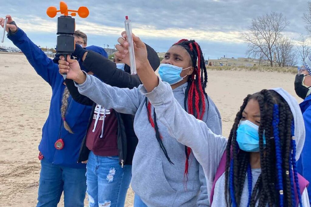 Four students stand on a Lake Michigan beach holding weather measuring instruments.