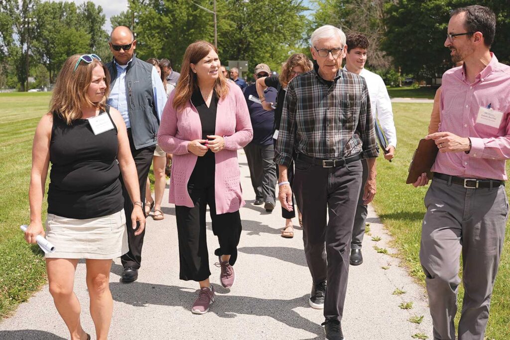 Julia Noordyk and Adam Bechle walk down a sidewalk while speaking with Kathy Blumenfeld and Governor Tony Evers.
