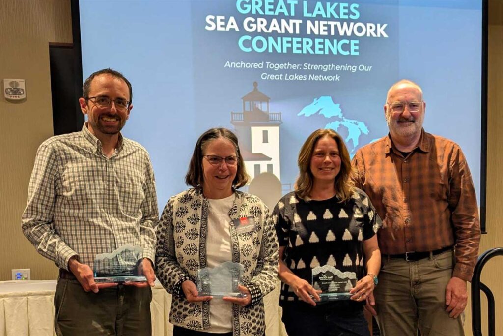 Adam Bechle, Ginny Carlton, Julia Noordyk, and David Hart pose for a group photo on the stage in front of a screen graphic that reads "Great Lakes Sea Grant Network Conference, Anchored Together: Strengthening Our Great Lakes Network."